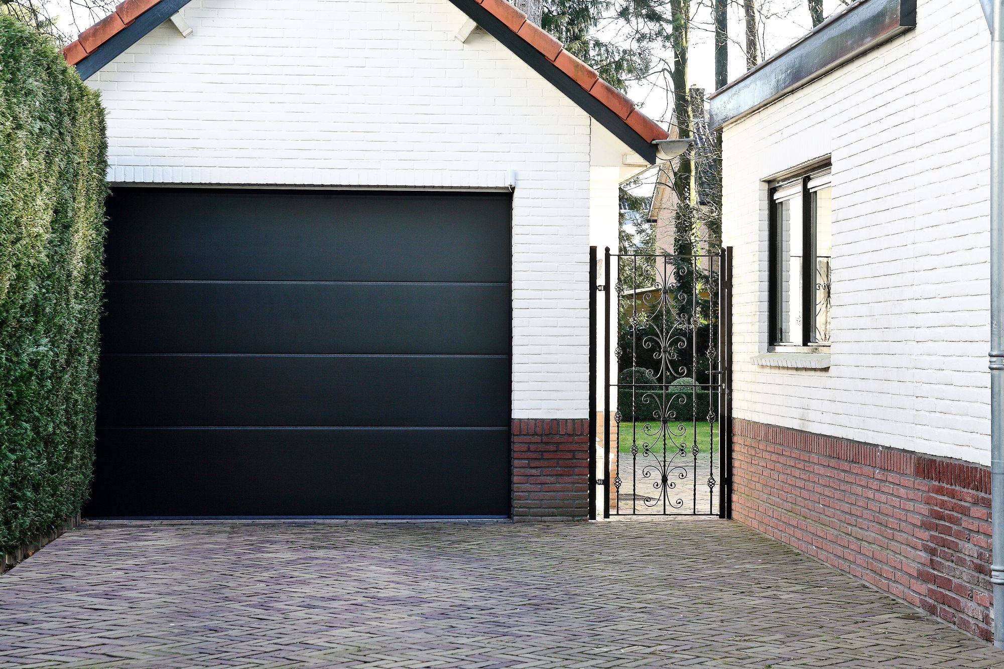 Zwarte sectionaal garagedeur naast een witte woning met smeedijzeren poort