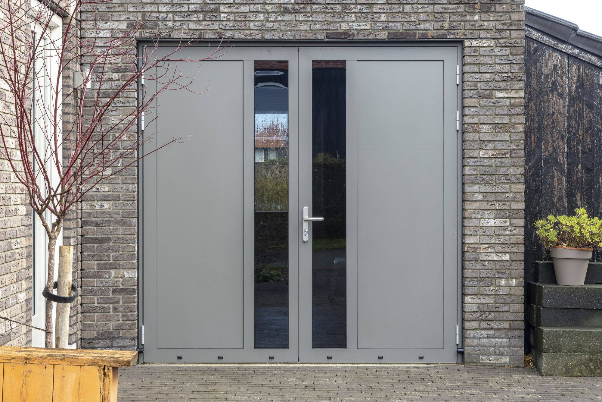 Grijze openslaande garagedeur met verticale glasstroken naast een houten bank